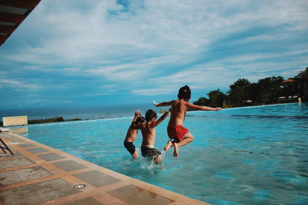 Three young children jump into a large pool on their family vacation, however, in the distance is a sign that states "off limits to swimmers". This image puts emphasis on why parents like you need travel insurance while travelling with kids as a Canadian family.