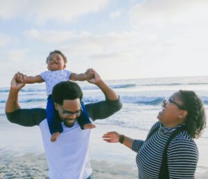 a family walks along the beach on their family vacation, their child toddler sits on his father's shoulders. The family is smiling, care-free and happy, because they got travel insurance for travelling with their kids.